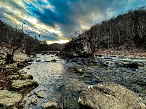 Bounder and rocks in a river lined with winterized trees at sunset