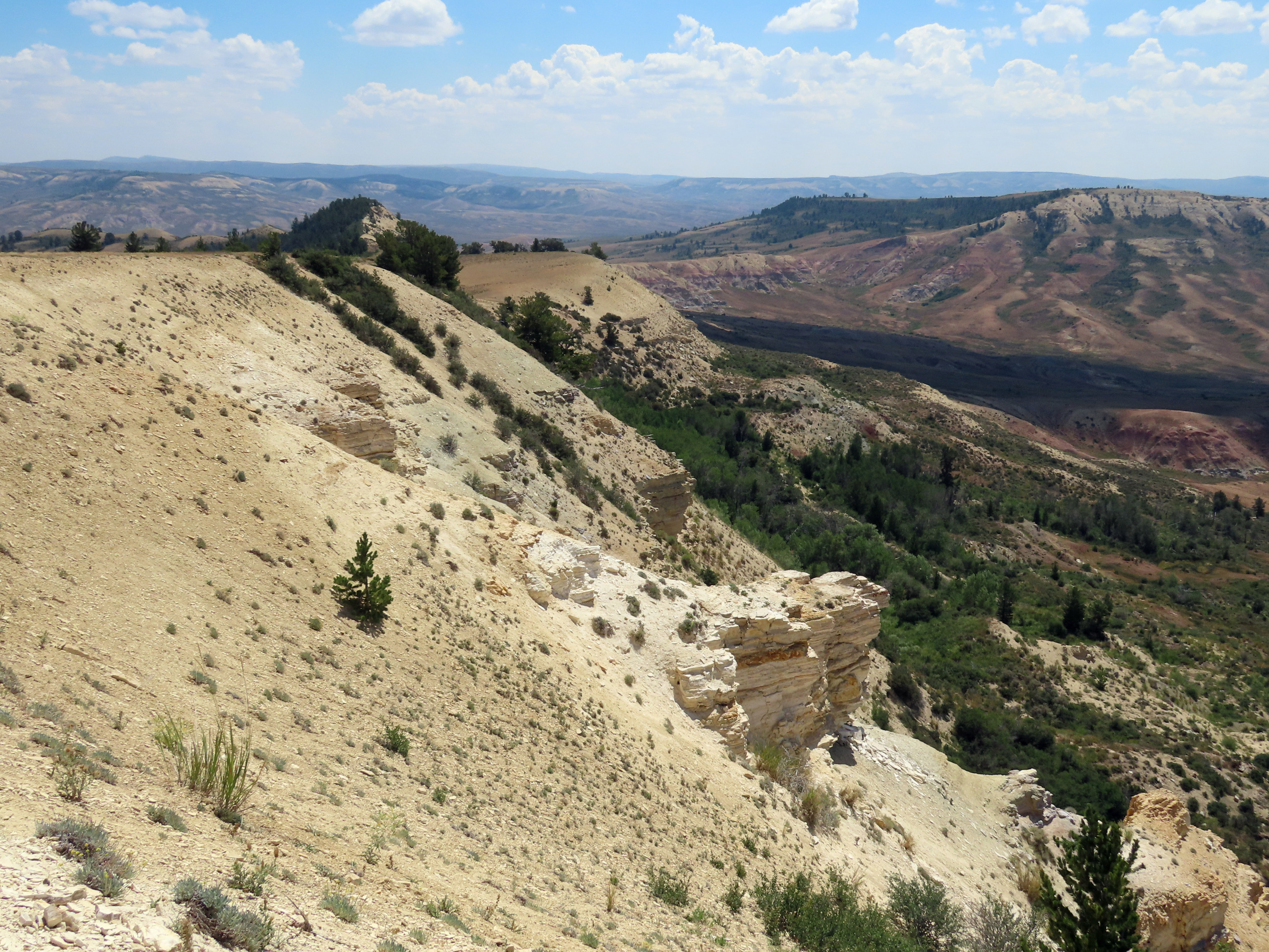 A ridgeline with craggy outcrops covered with some trees and shrubs sloping steeply down to valleys. There is another ridgeline visible on the other side of a valley.