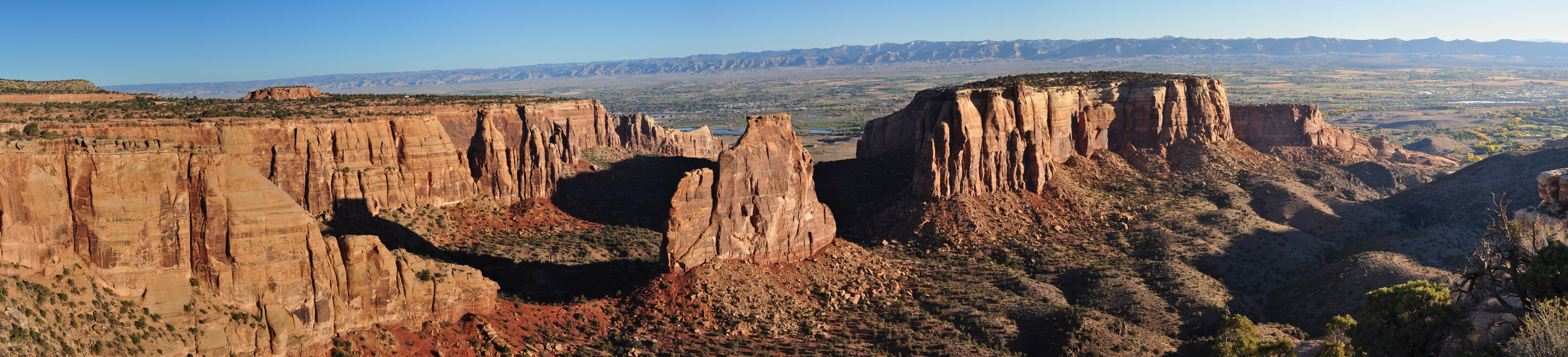 panorama of vertical reddish canyon walls from left to right broken by monolith and then rolling green canyon bottoms in shadow at right