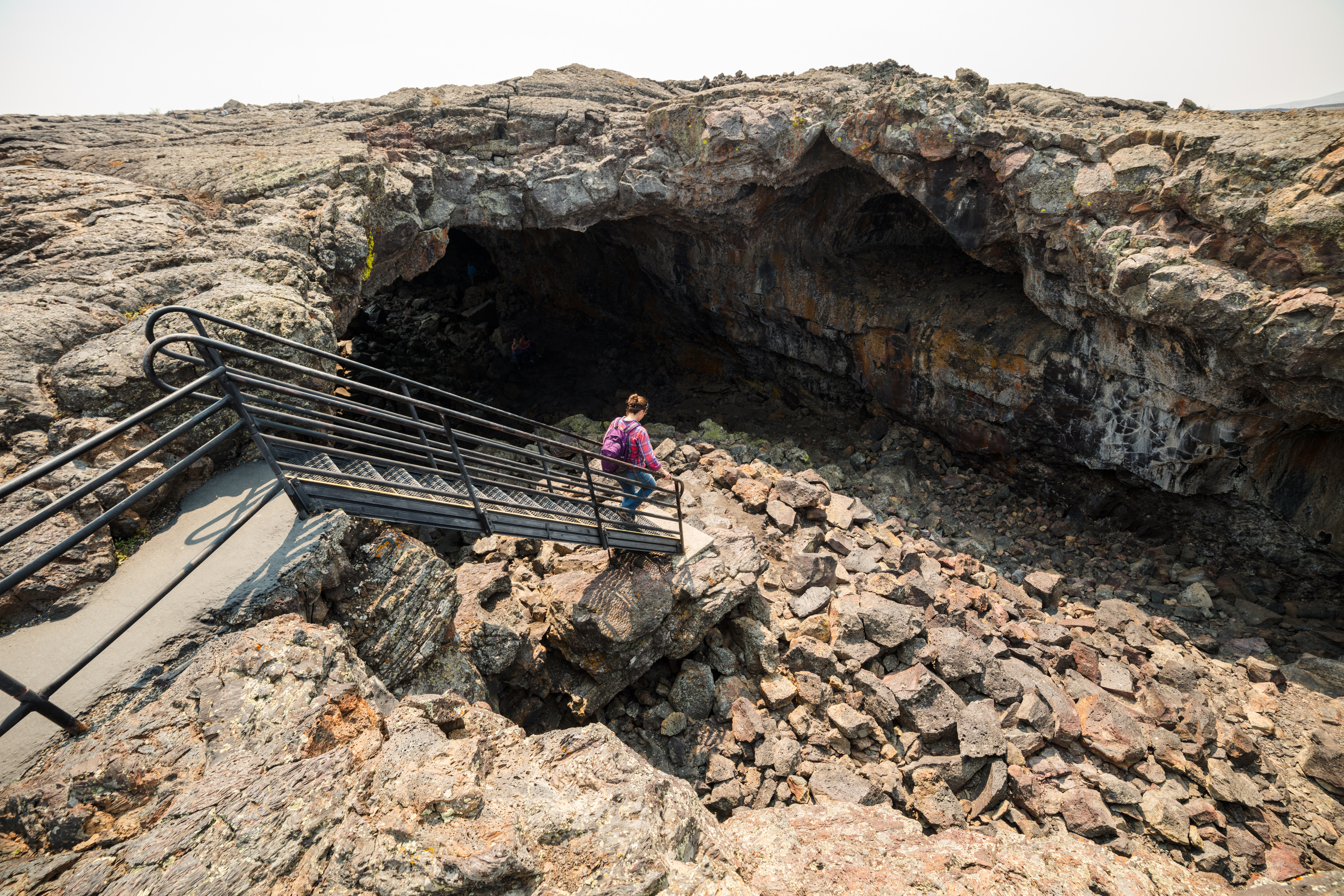 a woman with a backpack walking down a staircase into a large cave opening lined with many large, broken rocks