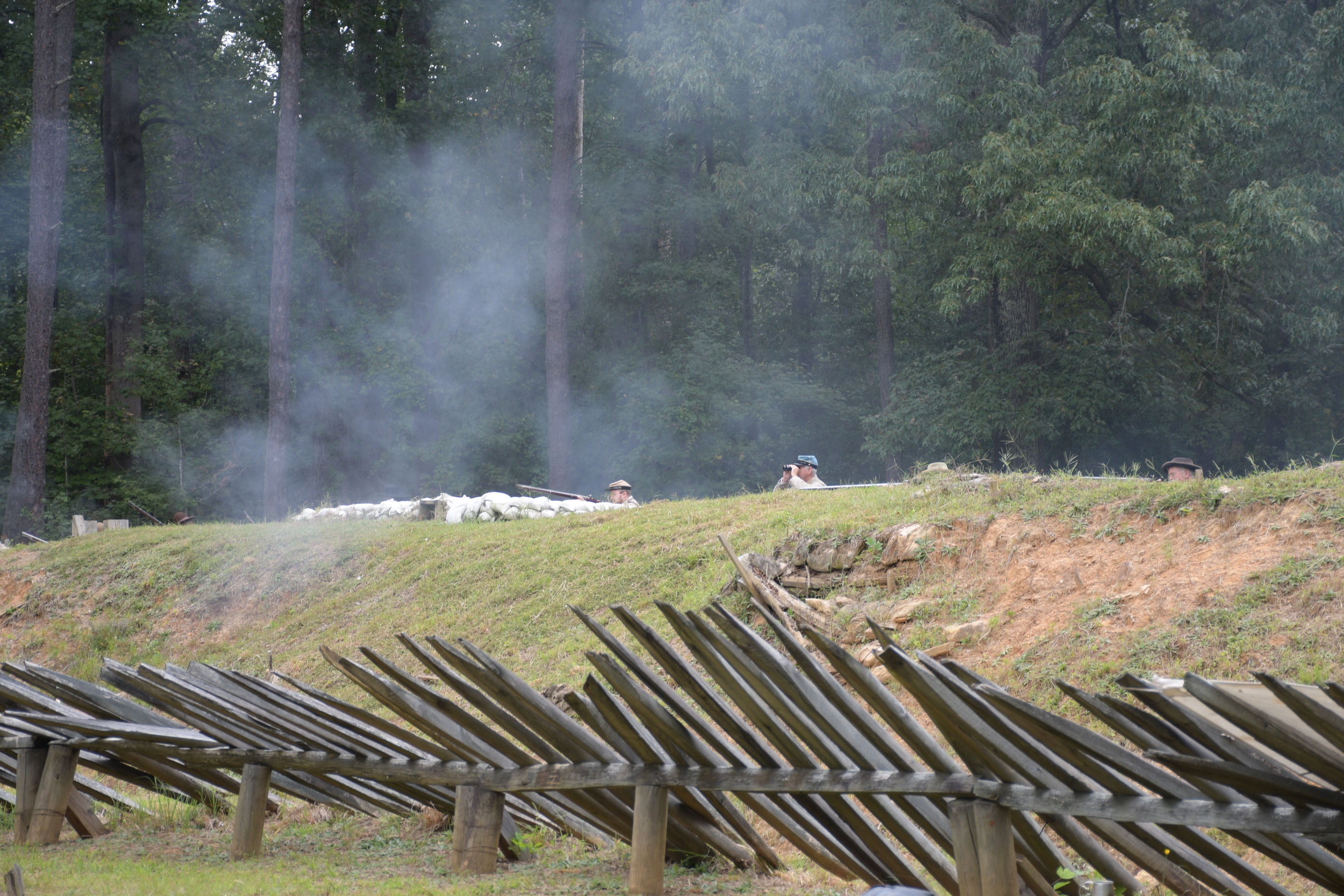 The heads of four white men wearing Confederate uniforms are visible above earthworks. One man is using a pair of binoculars the other three are firing rifles.