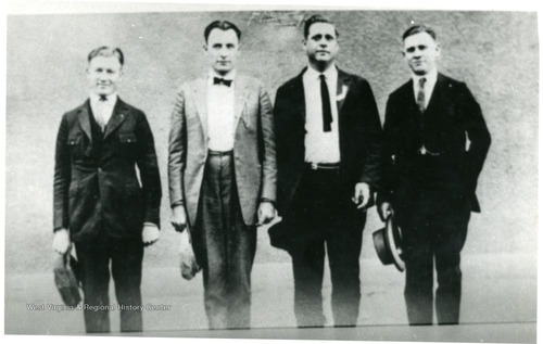 Six men in suits pose in a line in front of a brick building. Four are holding wide-brimmed hats in their hands. 