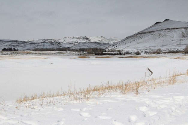 Snow covers a frozen body of water surrounded by mesas. The shoreline has yellow-brown grass and snow with footprints.