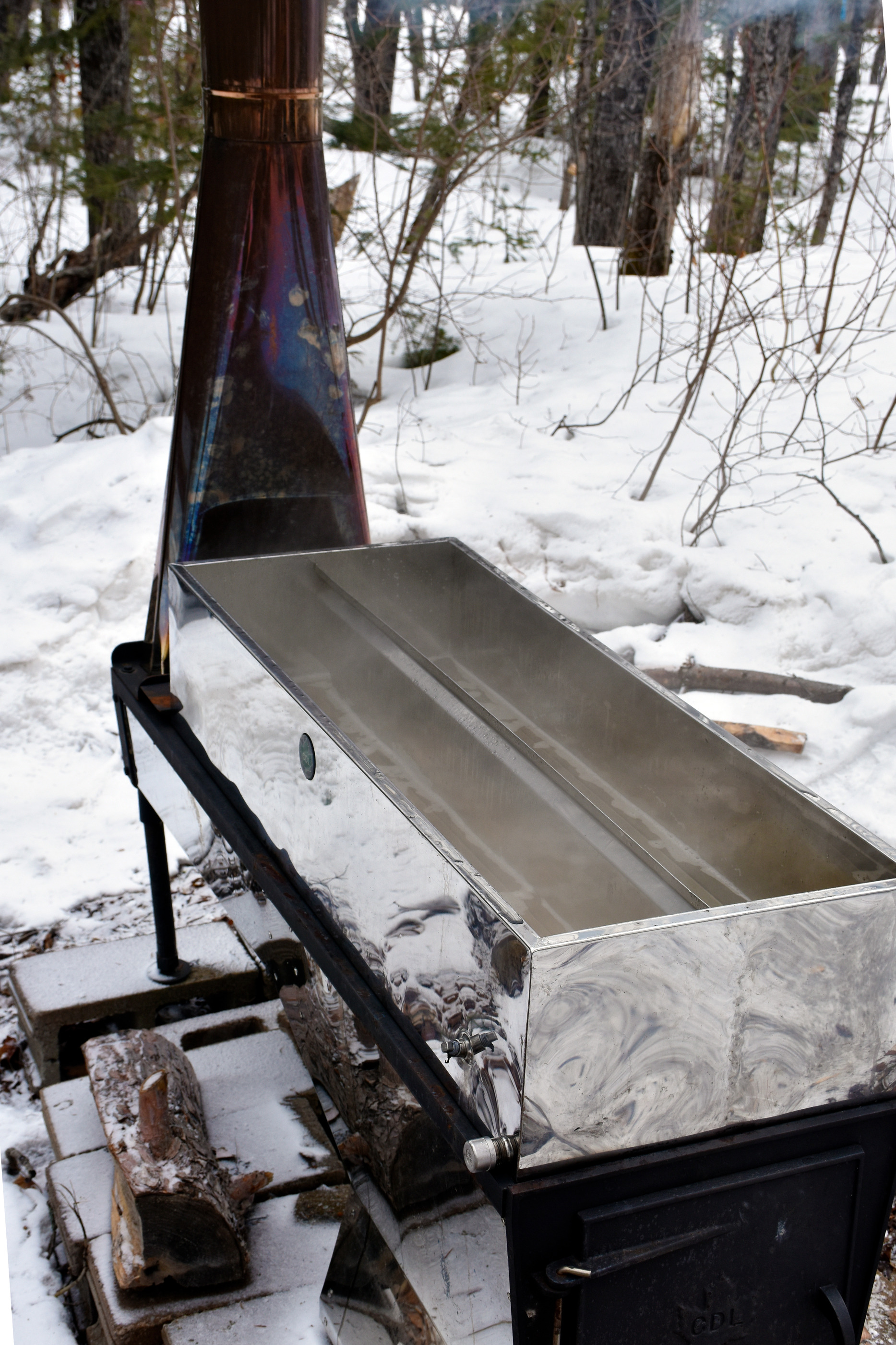 A stainless steel box with a divider in the middle steams from clear liquid boiling. A metal chimney draws smoke from the fire under the box (unseen).