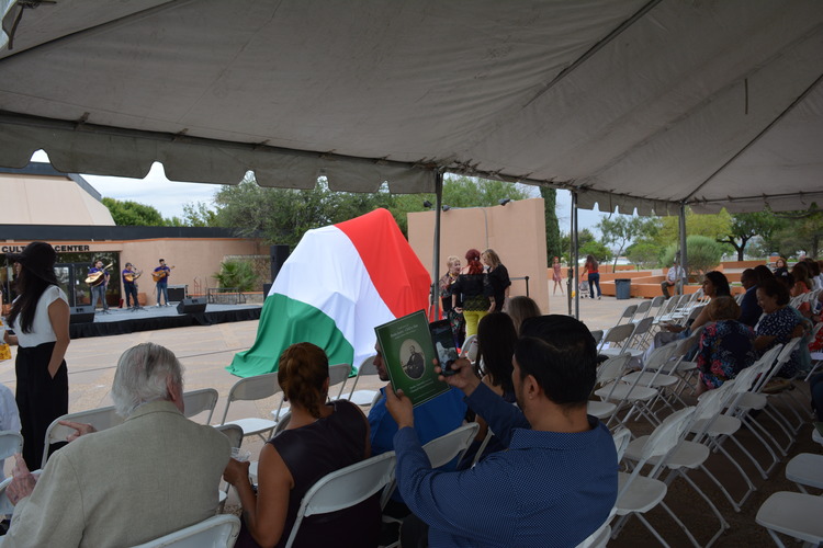 Rows of chairs under a shade canopy are partially filled with people. A green, white, and red cloth is draped over a large object just outside the tent. Behind that is a temporary stage with people playing stringed instruments.