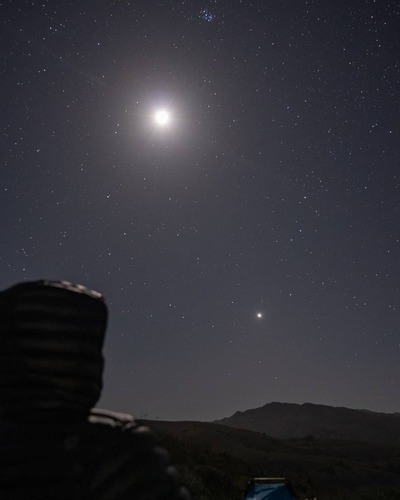 A bright white orb shines against a blanket of smaller stars on a dark night. In the left hand corner is a dark outline of a person in a hooded jacket sitting down and looking at the sky.