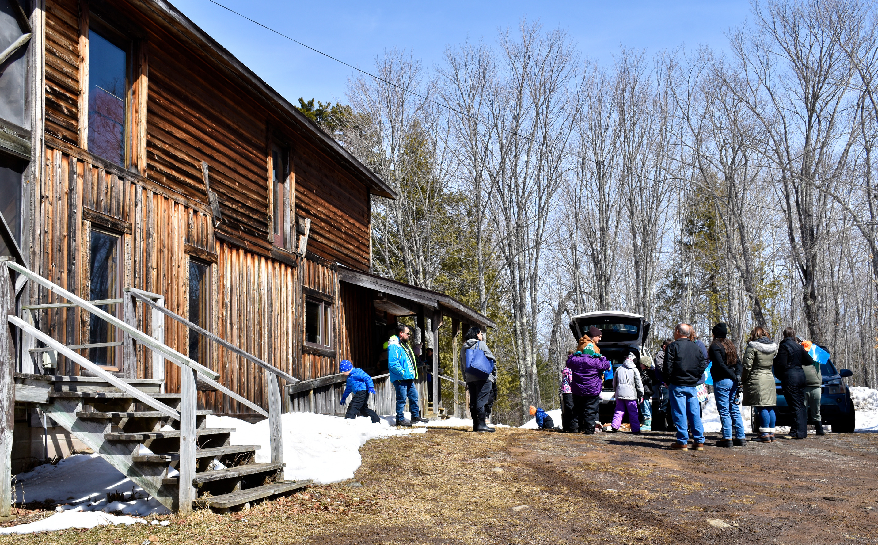 A large wooden building borders a dirt parking area where people are assembled near a car.
