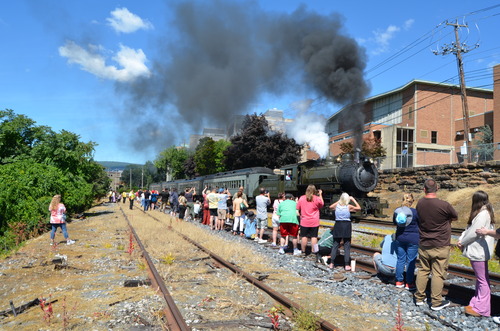 Steam locomotive puffing smoke and pulling a line of passenger coaches zips by a crowd of onlookers and photographers. 
