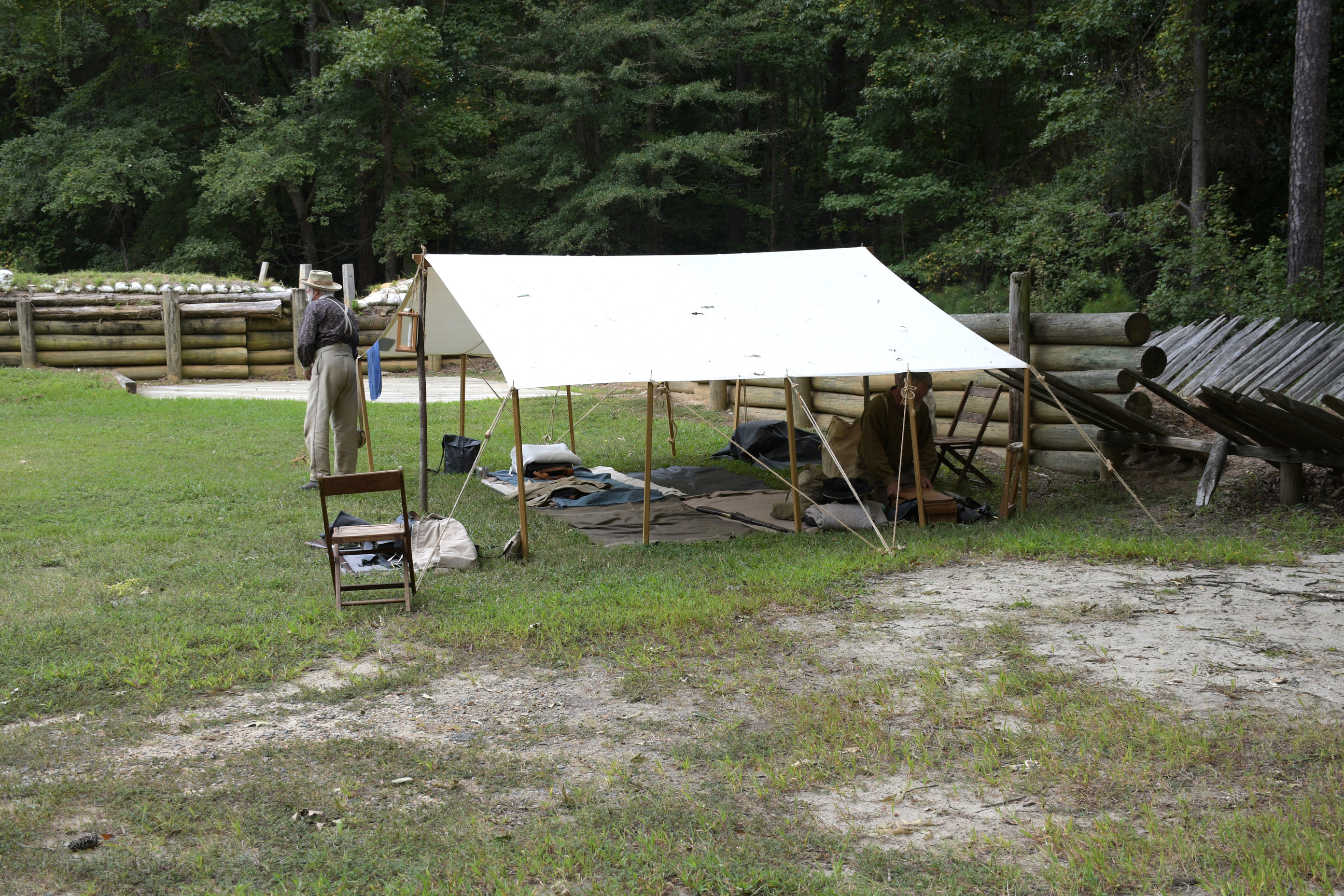 Two white men in confederate uniform and equipment and sleeping gear are in a large a-frame tent with no sides. Wooden palisade and wooden retaining wall supporting earthworks in the background.