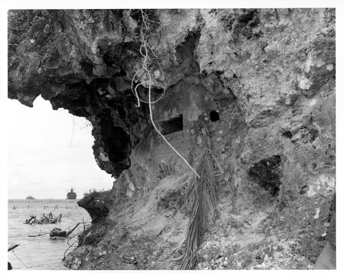 A man-made rectangular opening in the side of a rocky cliff. A palm frond and other vegetation conceals the hole. In the background is the ocean, full of barbed wire and other obstacles. Two battleships float in the background.