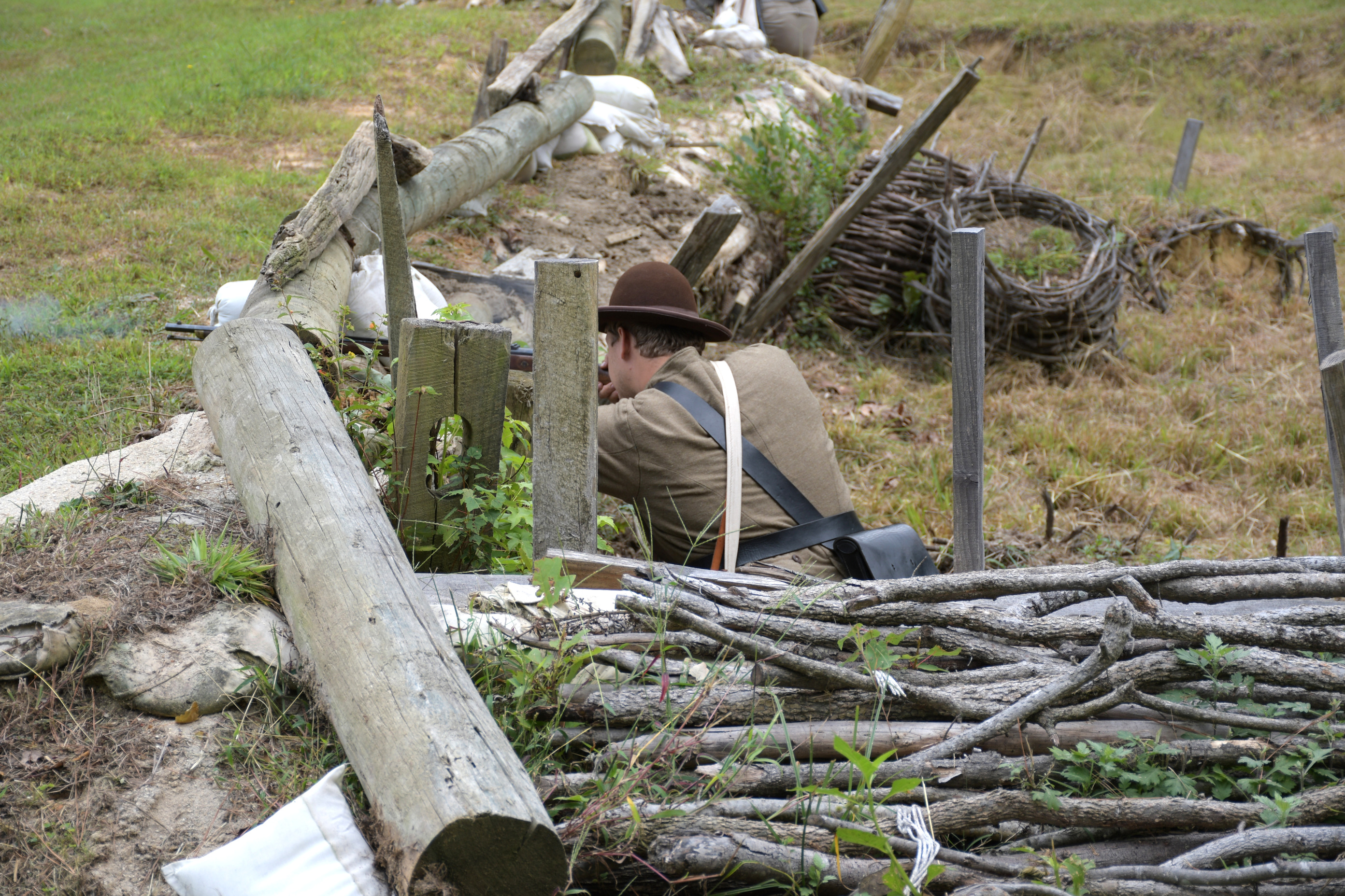 The back of a white man wearing a Confederate uniform and brown hat kneels behind earthworks - fires a rifle.