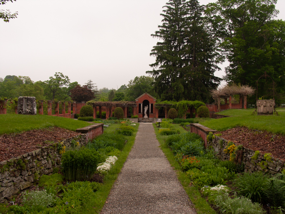 A long gravel path flanked by flowering shrubs and stone walls leading toward a pergola with marble statue.