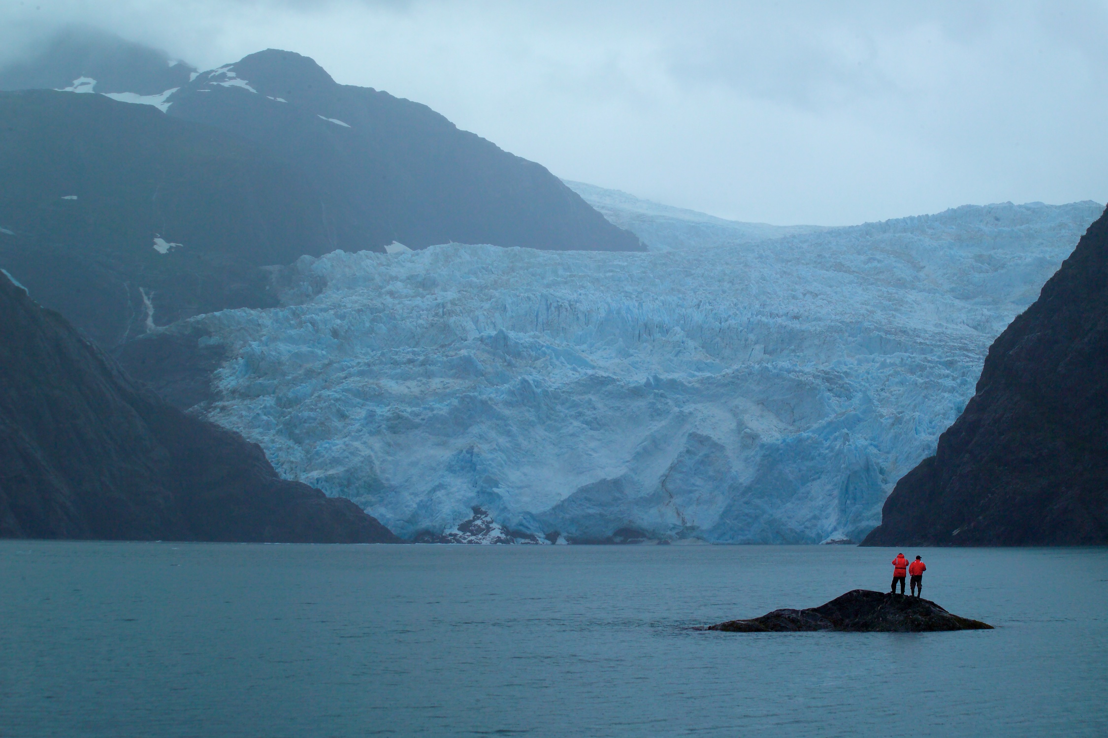 two orange  clad people stand on a small isle in a bay while looking at a icy wall nestled between cliff walls 