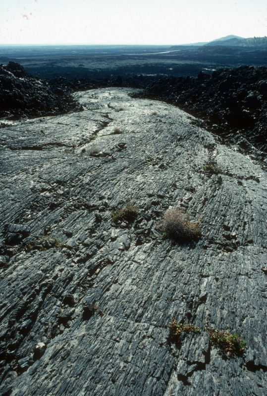 a smooth, wide ribbon of rock cutting across a field of black chunky rocks with a cinder cone and road in the distance