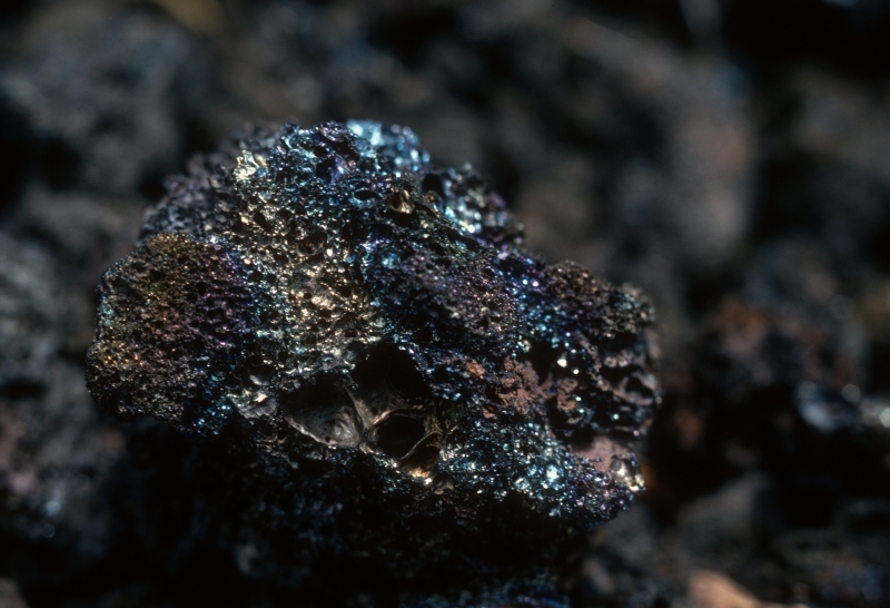 a closeup of a small black rock with many air holes and a shiny multicolored glaze 