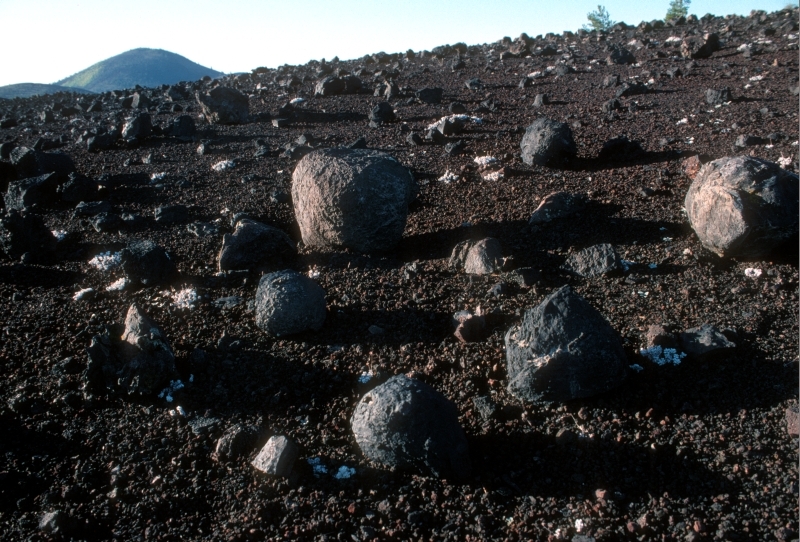 a field of dark gray rocks of varying shapes and sizes on a black cinder hillside with a large cinder cone in the distance 