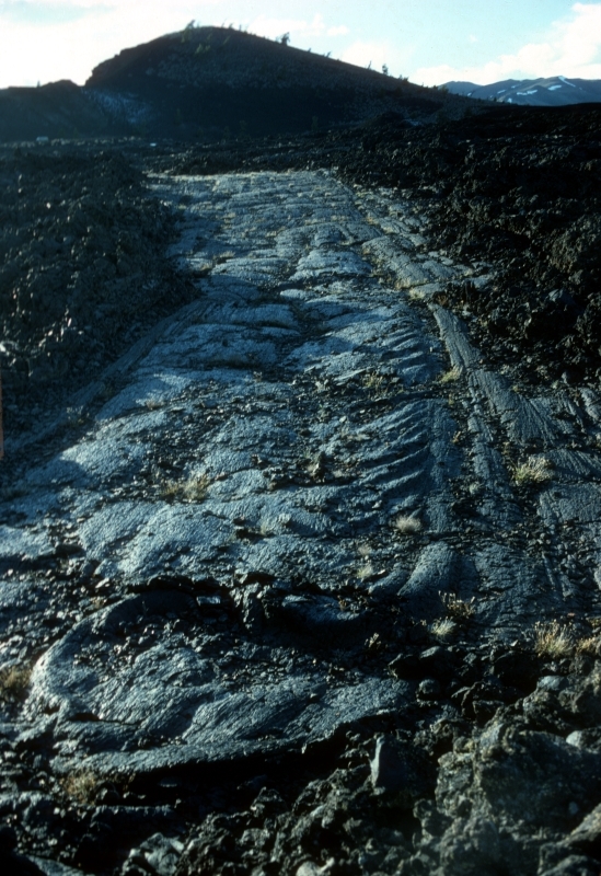 a smooth, wide ribbon of rock cutting across a field of black chunky rocks with a dark cinder cone in the distance