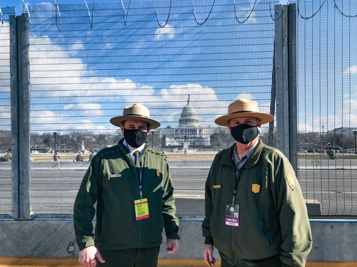 Two National Park Service employees standing next to a fence with the U.S. Capitol building in the background 