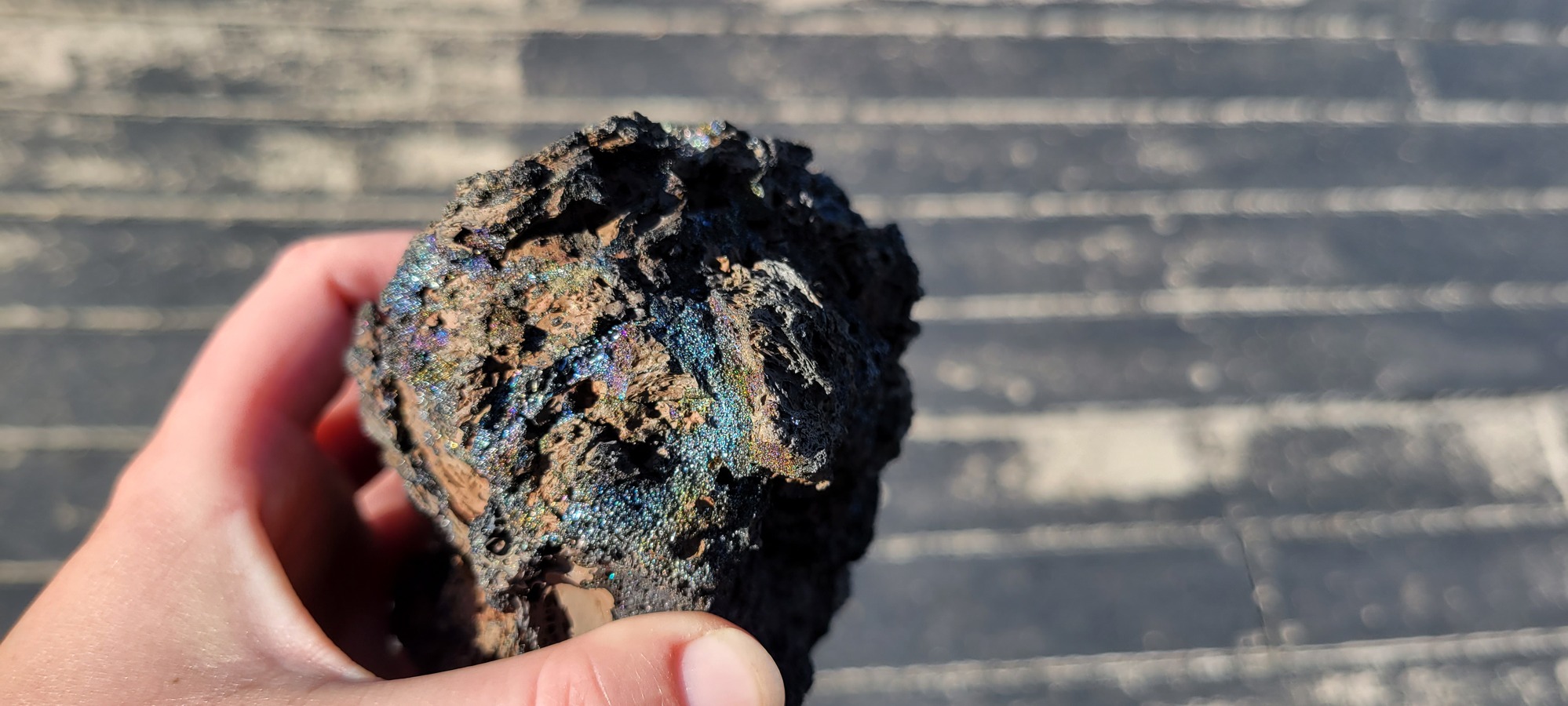 a rock about the size of the hand holding with many bands of a shiny blue and rainbow-colored glaze