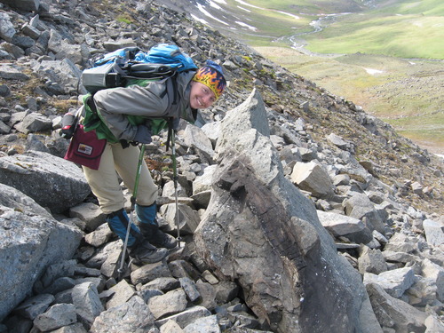 a young woman poses next to a rock with a log in it located in the side of rocky cliff with a green valley below.