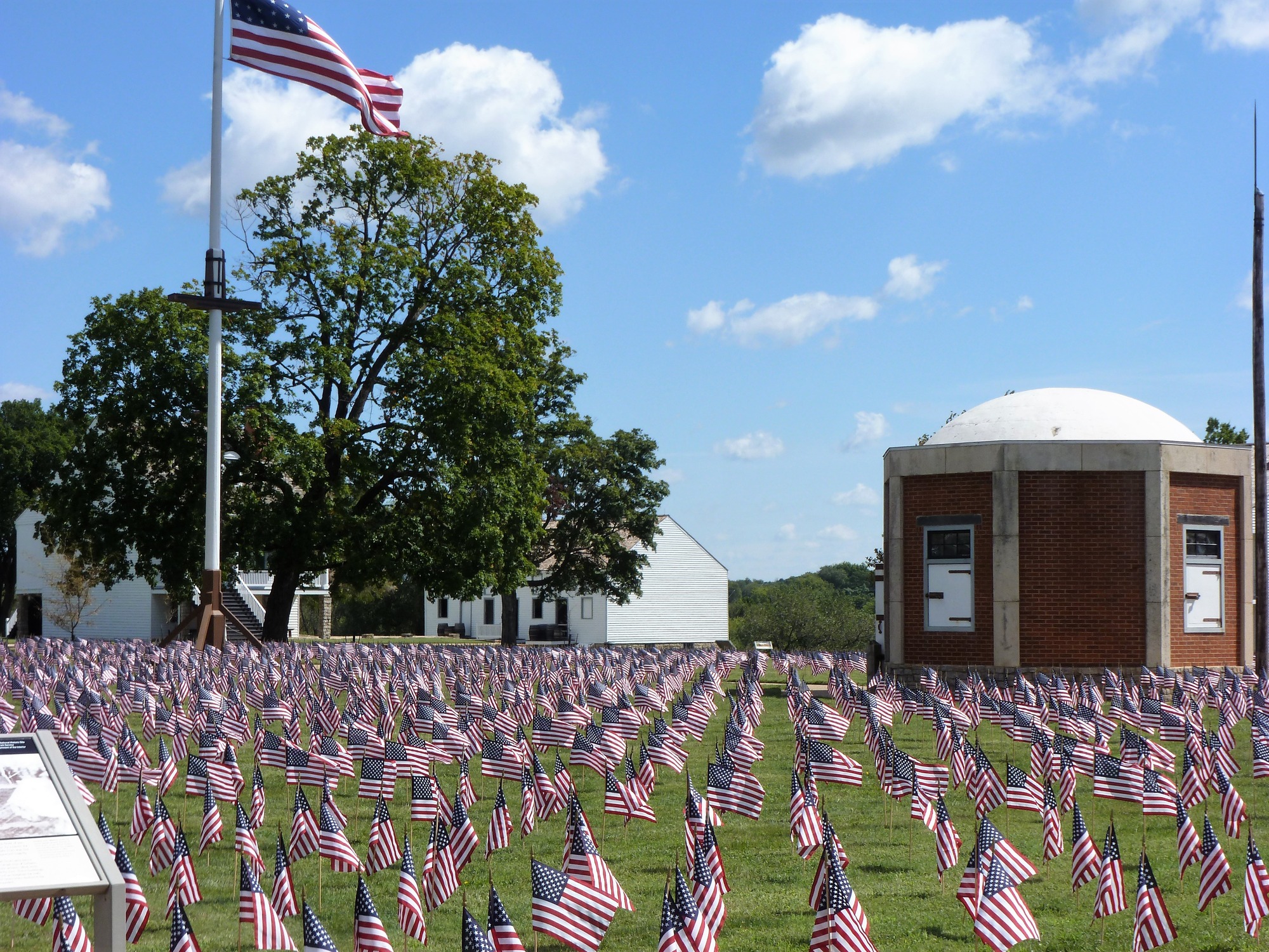 Small American flags cover a green field. A brick building with a domed roof is on the right, and on the left is a large American flag on a tall flagpole. The sky is blue with light clouds. 