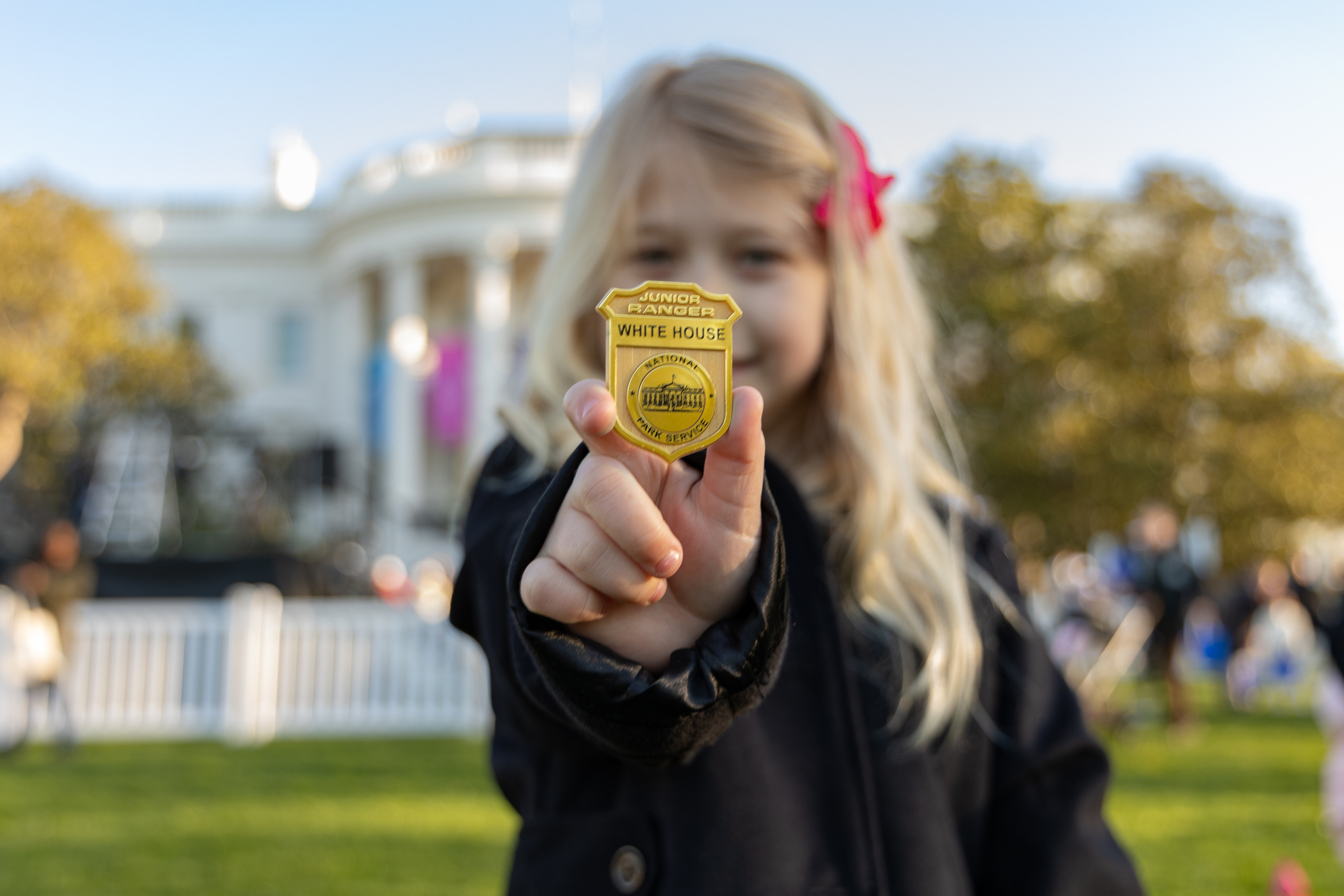 A junior ranger holds up a white house junior ranger badge in front of the white house