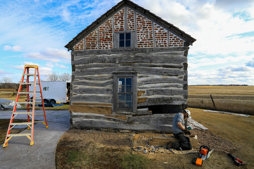 Log cabin at the base with red bricks filling the space under the roof. A man wearing hard hat, chaps, and gloves sits in front of the cabin on a log and cuts into a log using a mallet and chisel. A chainsaw is on the ground not far from his knees. An A-frame ladder is found on the sidewalk to the left of the cabin.