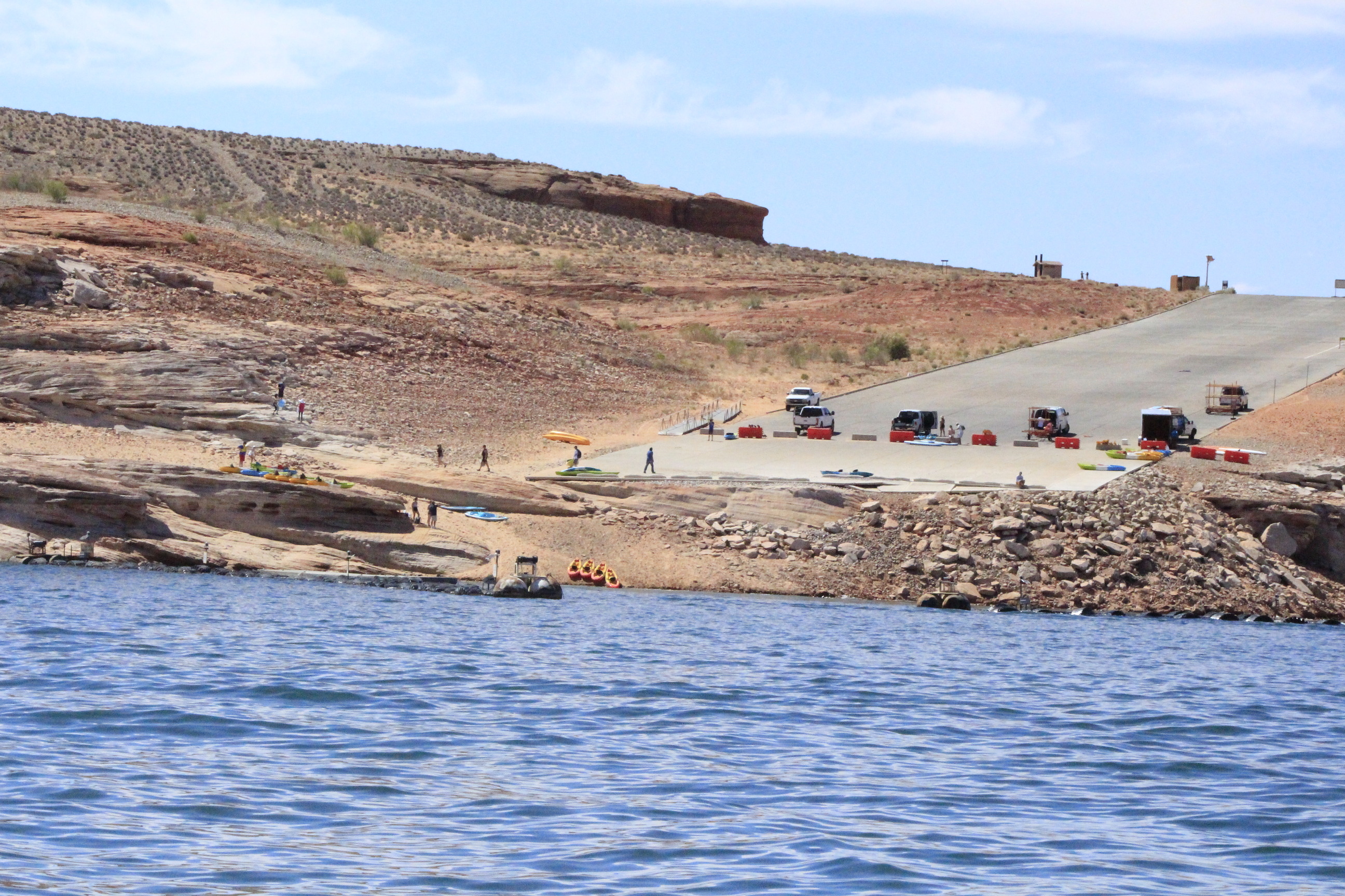 Trucks unloading kayaks on a barricaded launch ramp as visitors descend a rocky shoreline.