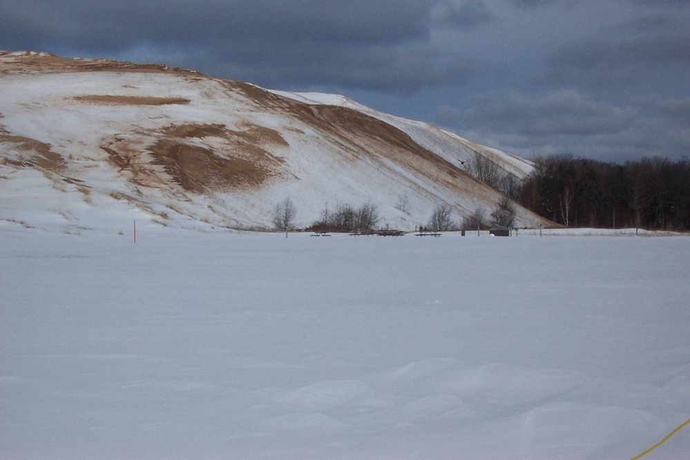 The Sleeping Bear Dune Climb in winter — fresh snow covers the dune face with bare trees silhouetted against a gray winter sky