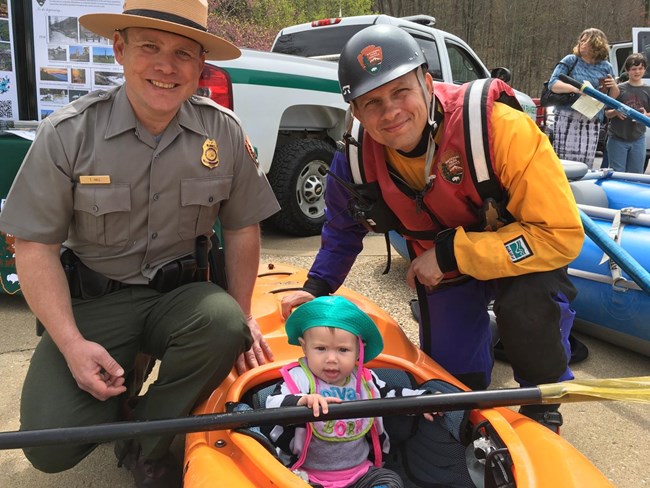 A young girl is sitting in a kayak. There are two park rangers sitting on either side of her. The ranger on her right is wearing whitewater paddling gear. The ranger on her left is wearing his park service uniform.