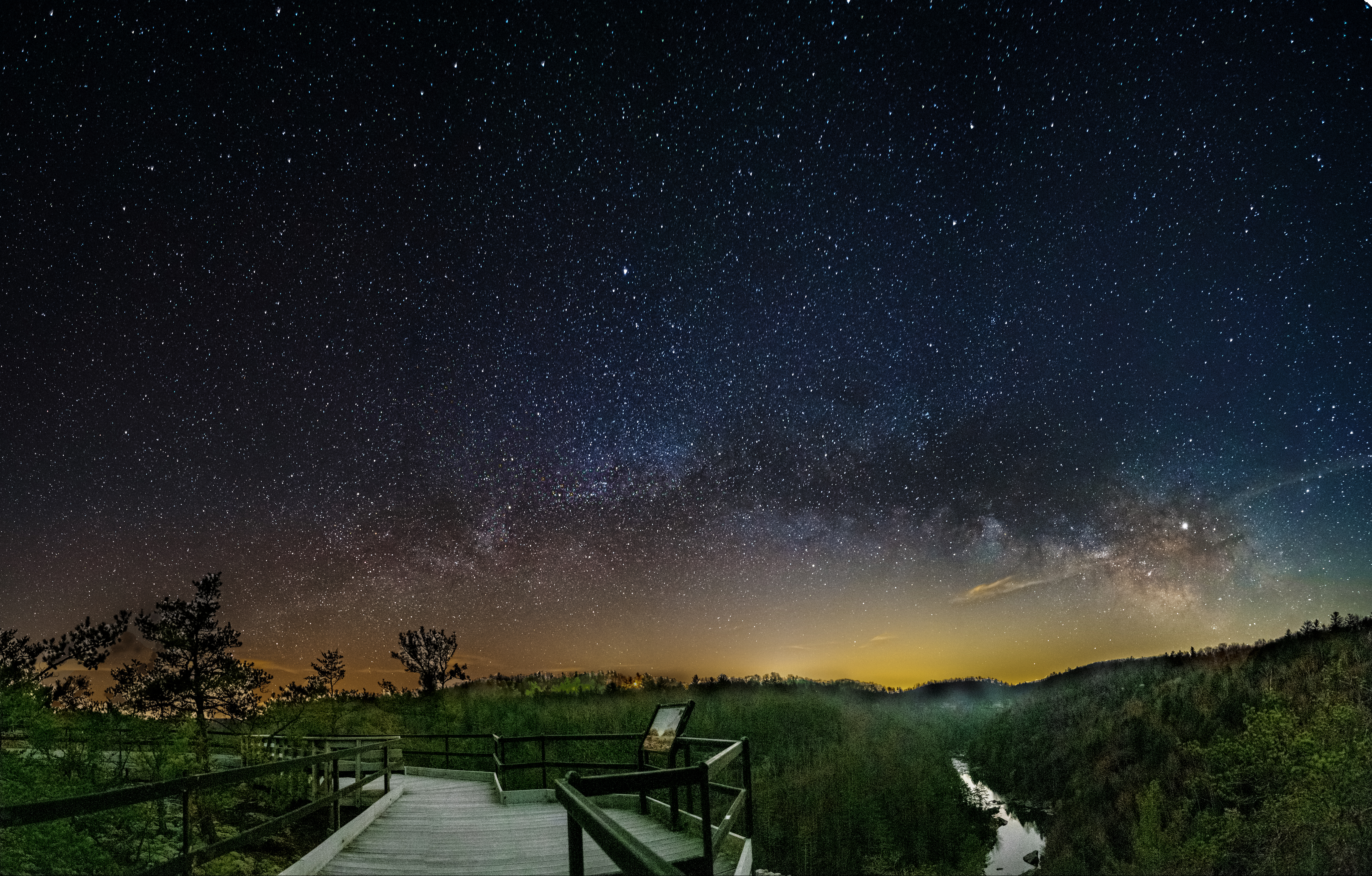Many stars illuminate the night sky over a forested area, river, and wooden overlook.