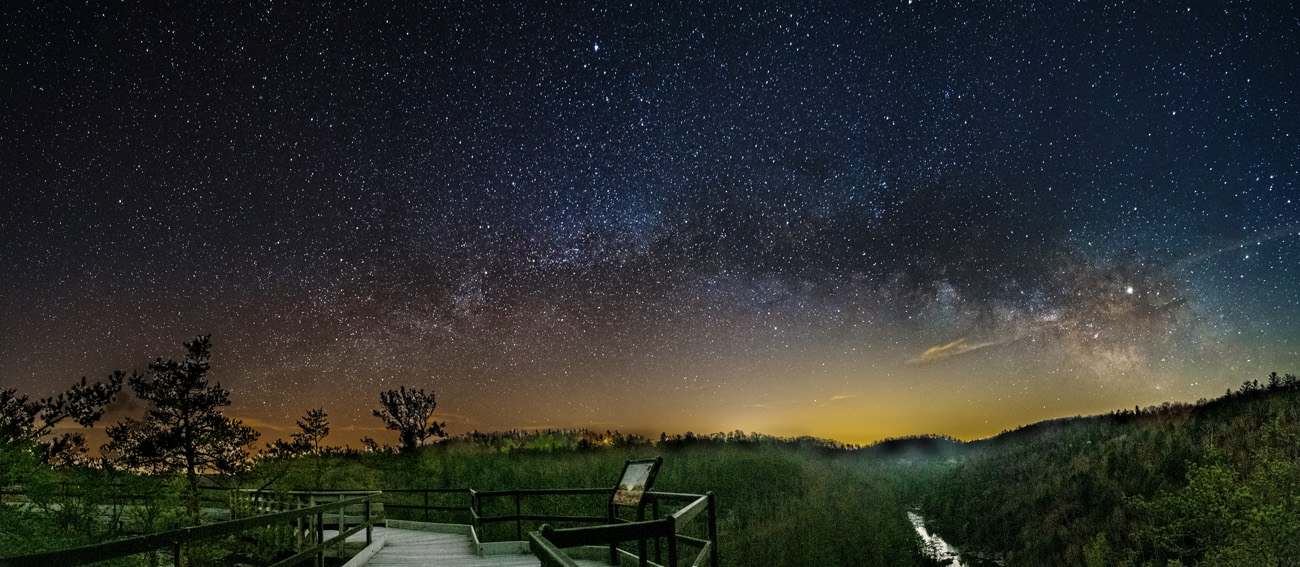 Many stars illuminate the night sky over a forested area, river, and wooden overlook.