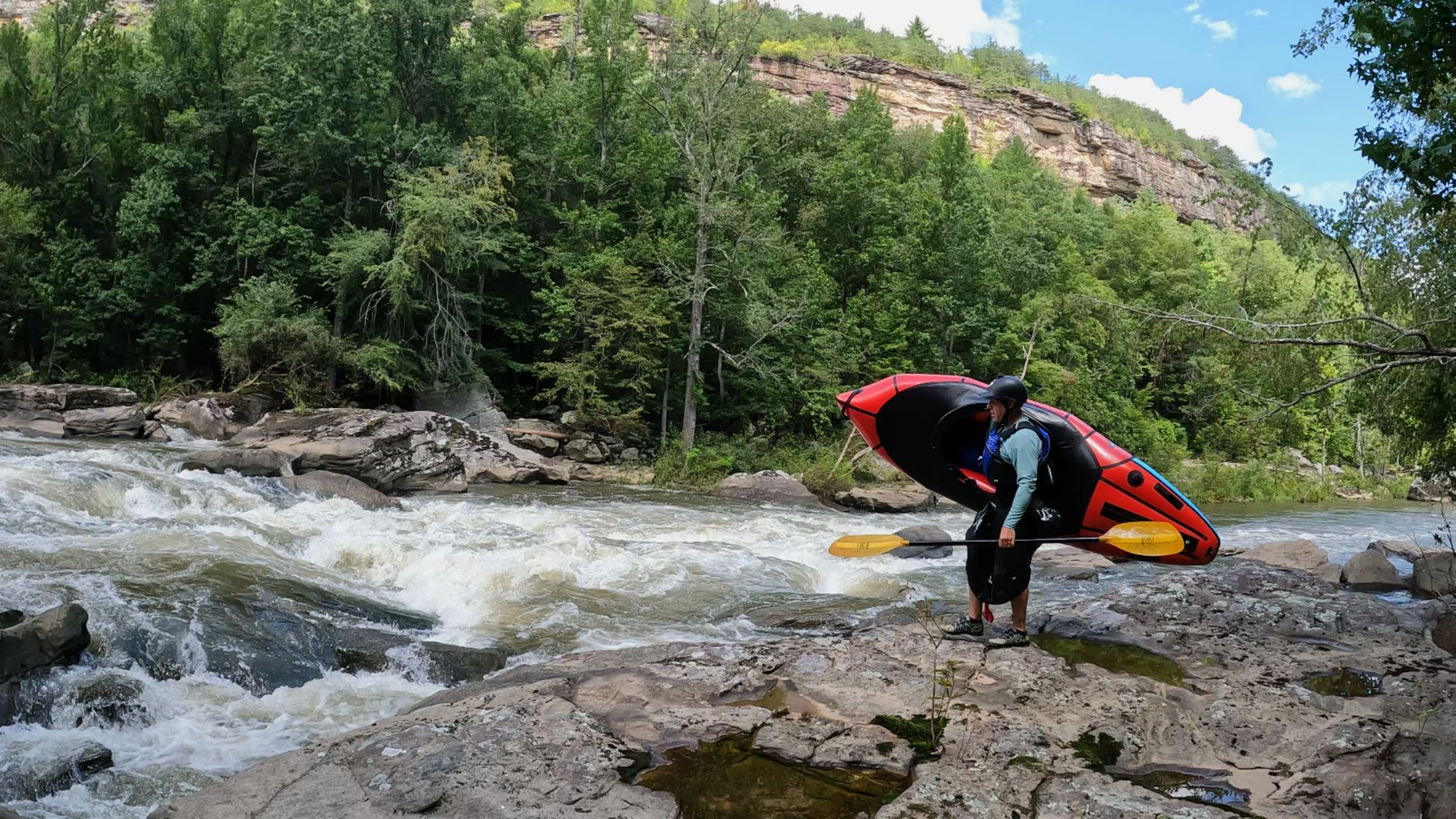 A kayaker stands on rocky terrain near a river, holding a kayak on their shoulders. The kayaker is facing the river, which is visible in the background. The scene depicts a moment of pause and contemplation.