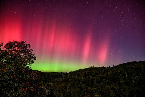 An aurora stretches across a starry night sky, an intense array of red, purple, and green. Small, forested mountains are seen in the bottom third of the photograph.