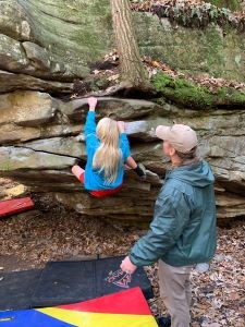 A young girl bouldering with a spotter.