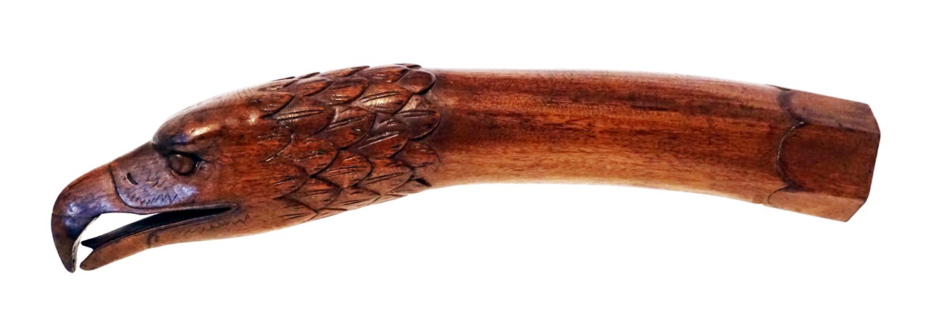 A wooden tiller. An eagle's head with its beak slightly open faces to the left. The right side is cylindrical shaped and the end is hexagonal.