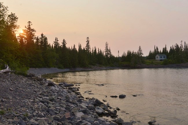 sun setting on the horizon over treeline with cabin on left and placid lake in the foreground
