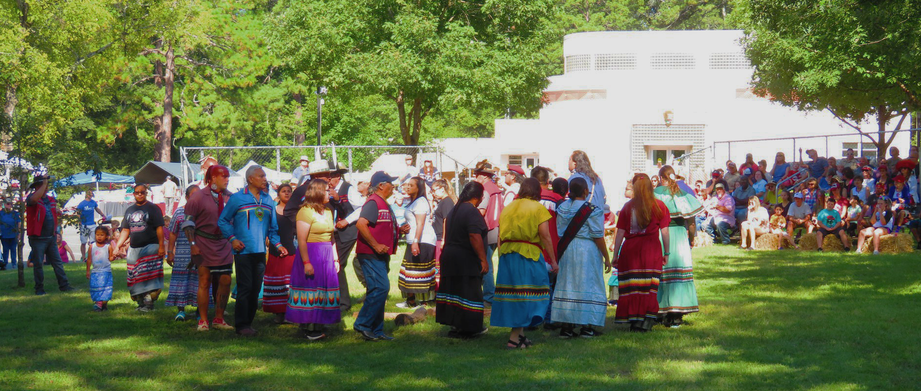 A group of Indigenous people performing a stomp dance in front an audience. The visitor center is in the background