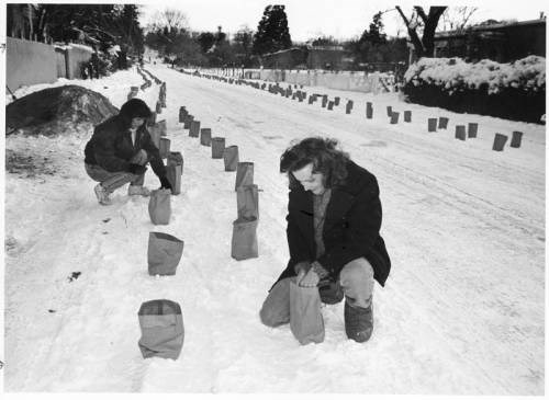 Two women prepare farolito lanterns along a snowy road. Canyon Road Acequia Madre 1990.