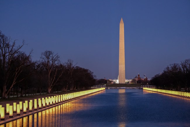 lt= At night, memorial lanterns line the reflecting pool across from the Washington Monument in Washington D.C.