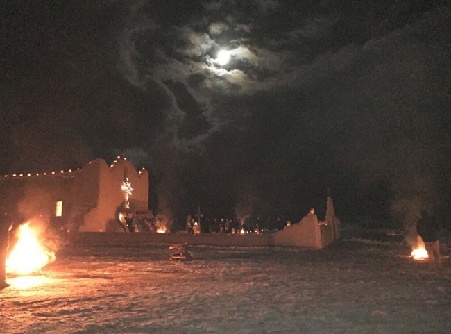 At night, luminarias and bonfires at Picuris Pueblo Church, an adobe building with a low adobe wall.