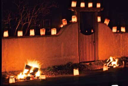 At night Paper farolito lanterns are lined up on top of an adobe wall. Two small bonfires are burning in the foreground.