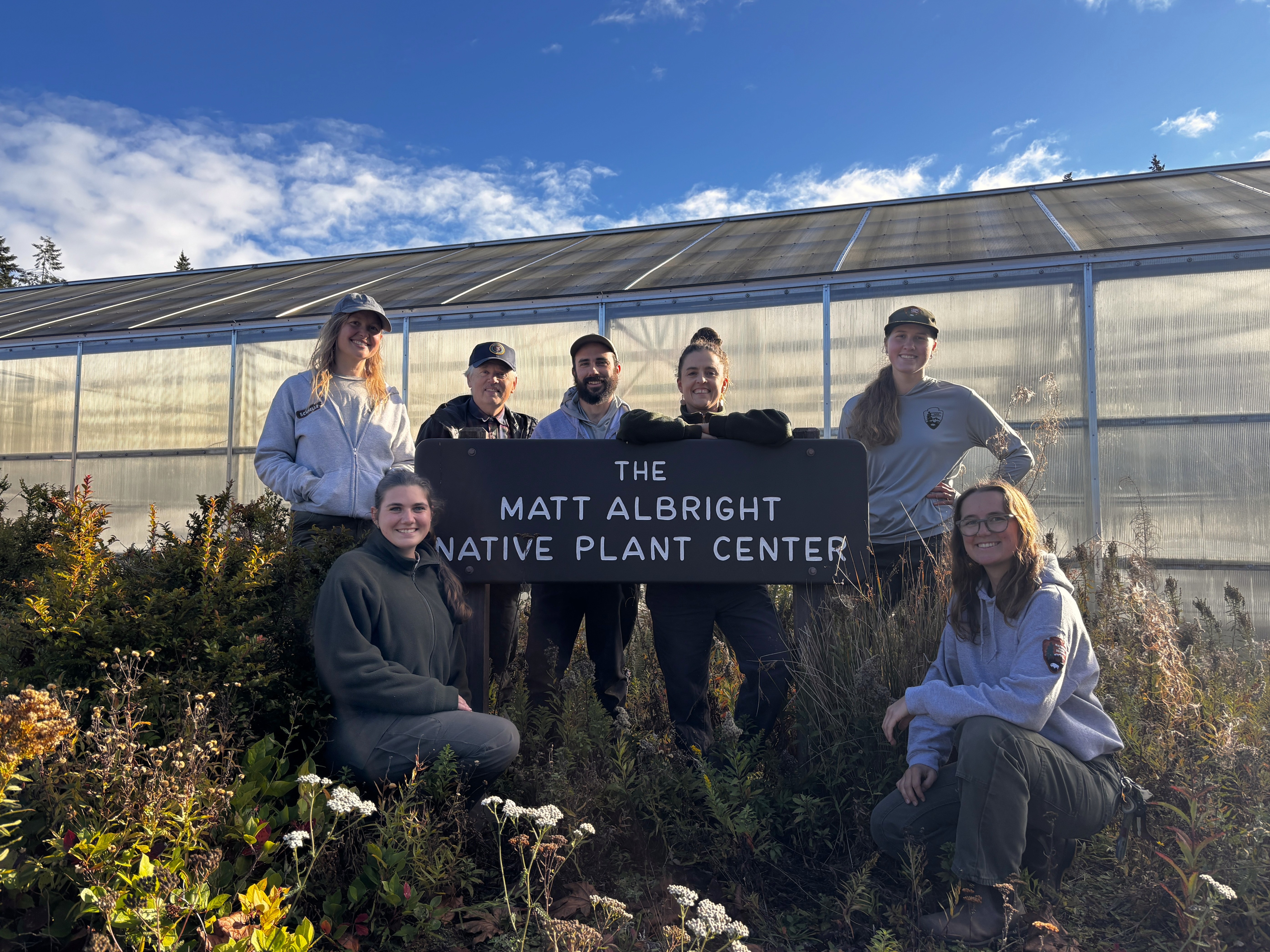 a group of people in baseball caps and hoodies gathered outside a greenhouse at the Matt Albright Native Plant Center.