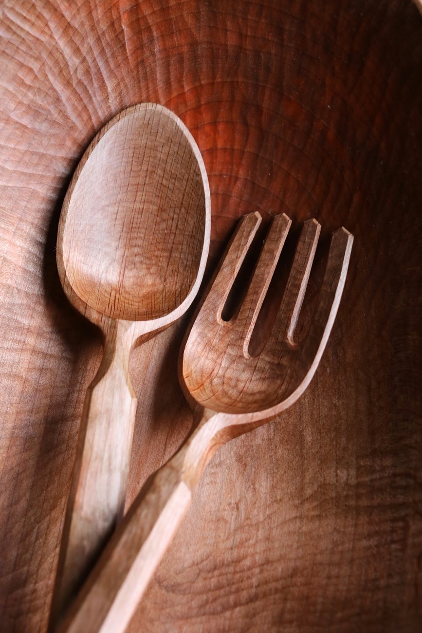 A carved, shining wooden fork and spoon inside a carved wooden bowl.