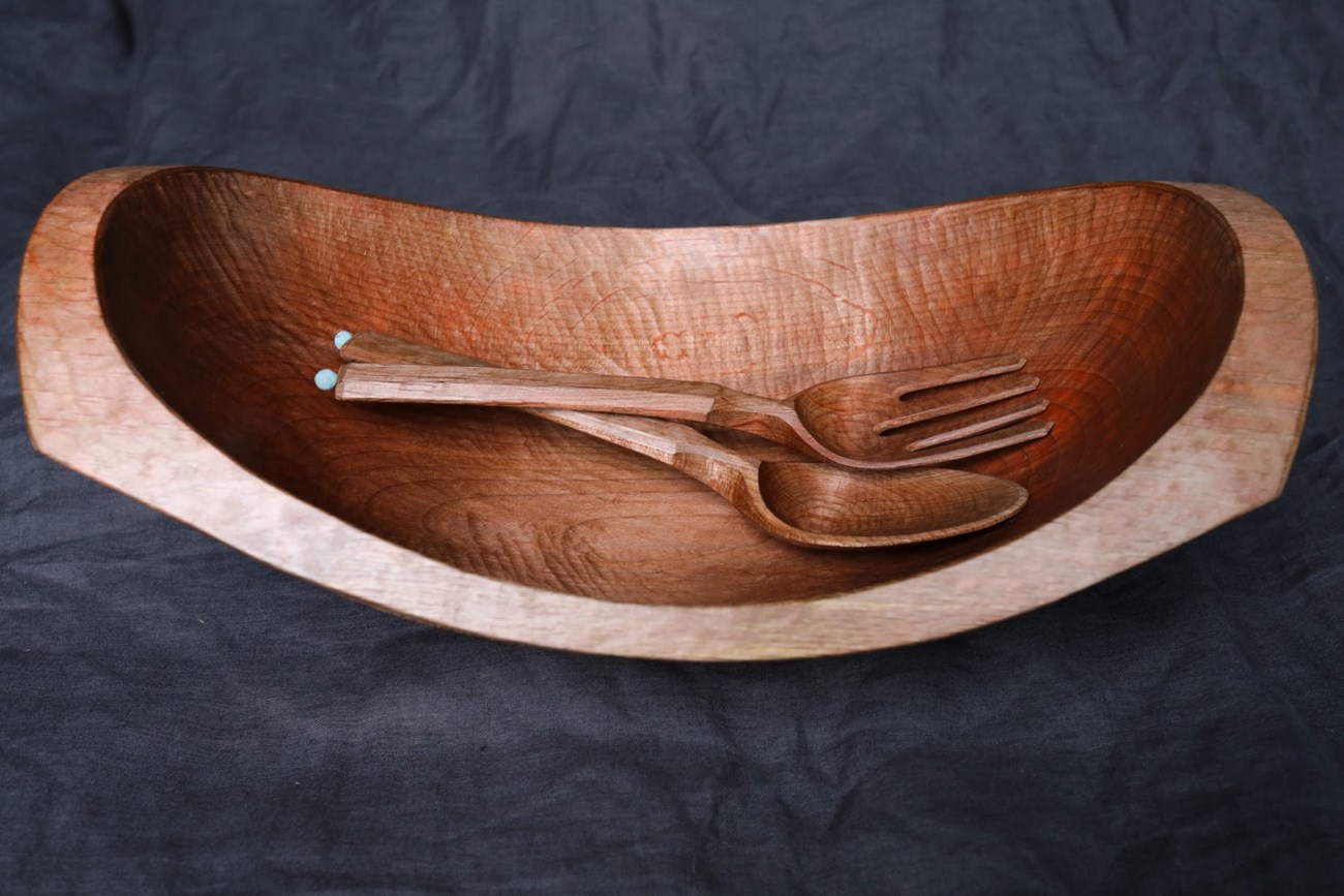 A carved, shining wooden bowl, wide and with a broad flat rim, with a wooden fork and spoon inside.