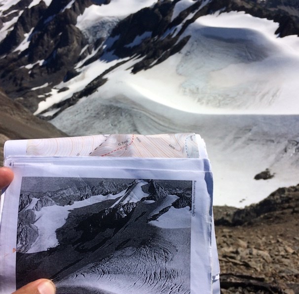A hand holds a black and white printed photo of a glacier, in front of the real glacier in the background.