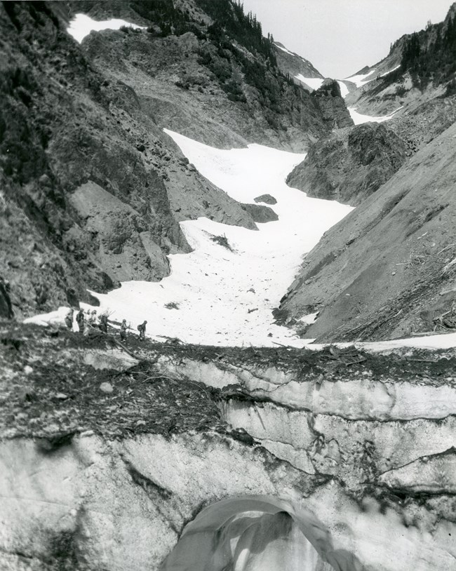 Archival photo, a group of people about to cross a snowy bridge at the foot of a long, narrow stretch of glacial ice.