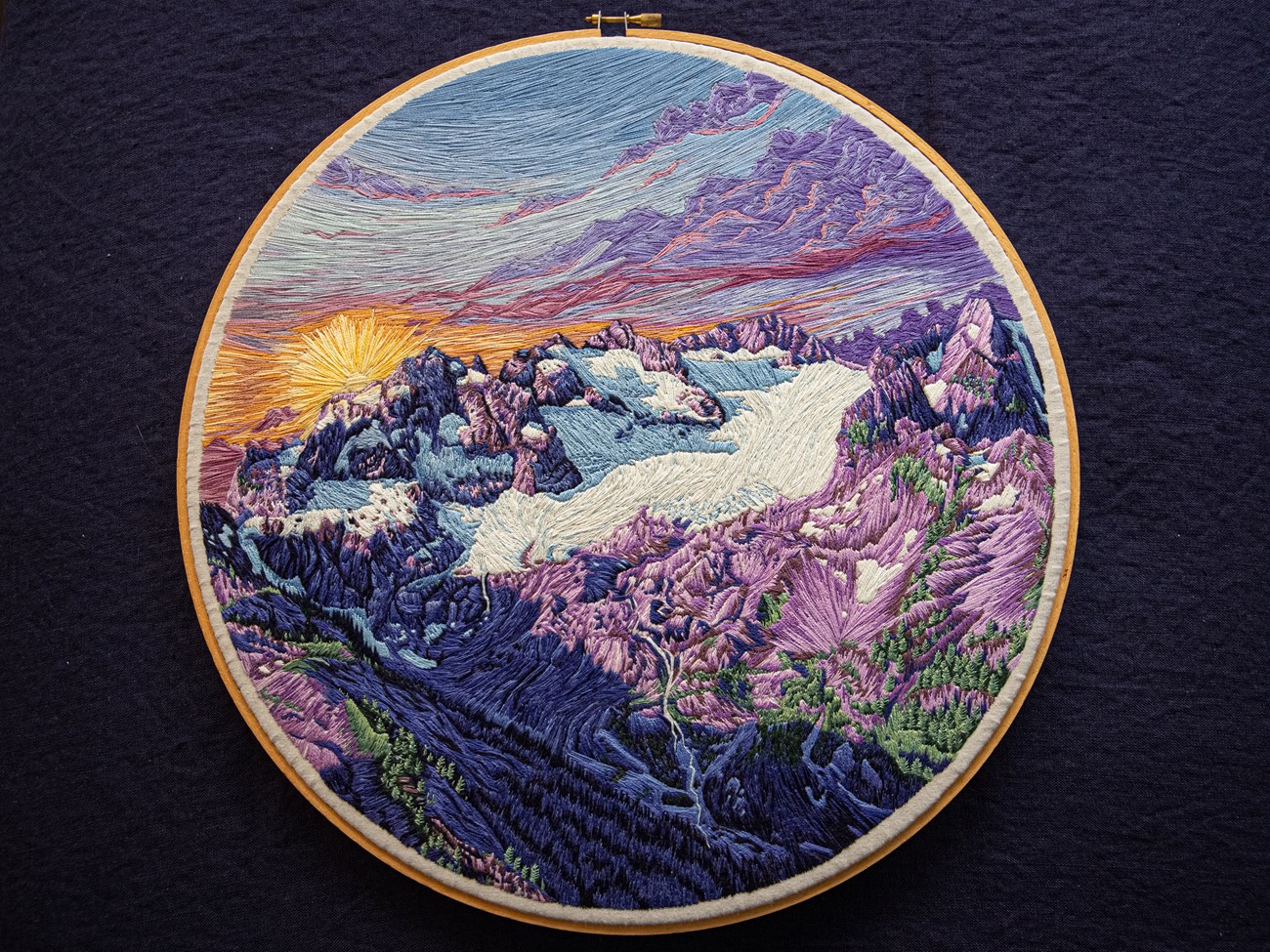 An embroidery hoop with a detailed sunset over a mountain glacier, rendered in threads of purple, white, gold, and blue.