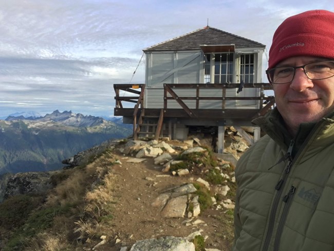 A man in a warm hat and insulated jacket stands in front of a mountain fire lookout.
