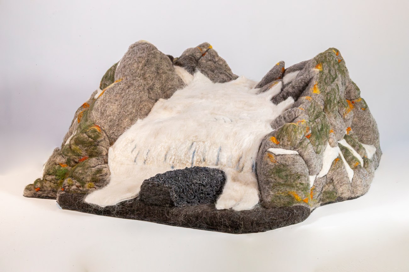 A mountain glacier sculpted from wool felt. The white ice of the glacier flows between grey “rock” ridges streaked with green and orange.